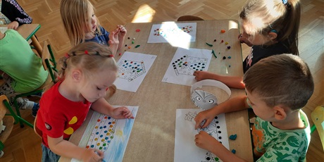 Powiększ grafikę: Czwórka dzieci siedzi przy stoliku i wykleja plasteliną obrazki.