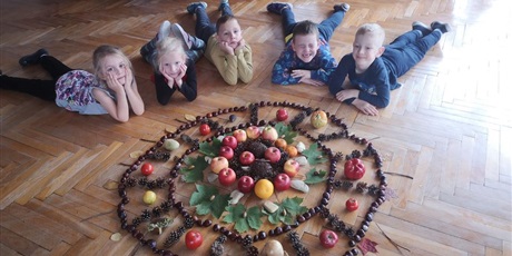 Powiększ grafikę: Pięcioro dzieci leży na podłodze przed mandalą stworzoną z owoców, warzyw, kasztanów, liści i szyszek.