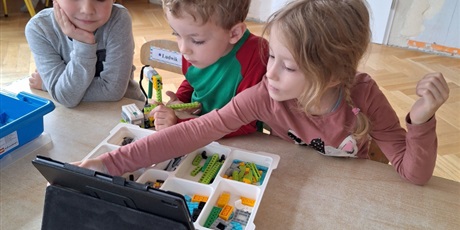 Powiększ grafikę: Dwóch chłopców i dziewczynka budują robota z klocków lego wg instrukcji na tablecie.