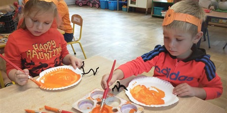 Powiększ grafikę: Dwoje dzieci maluje pomarańczową farbą papierowe talerzyki.