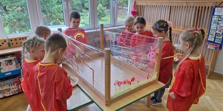 Powiększ grafikę: Dzieci malują farbami po foli spożywczej przyczpionej do nóg stołu odwróconego do góry nogami.