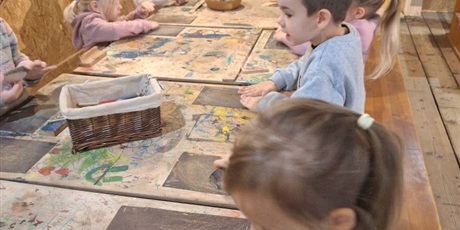 Powiększ grafikę: Kilkoro dzieci przy stole pociera kawałki drewna o papier ścierny przytwierdzony do stołu.